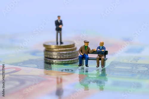 Miniaturgeschäftsmann auf Münzstapel hinter Rentnern auf Parkbank – Symbolbild soziale Ungleichheit