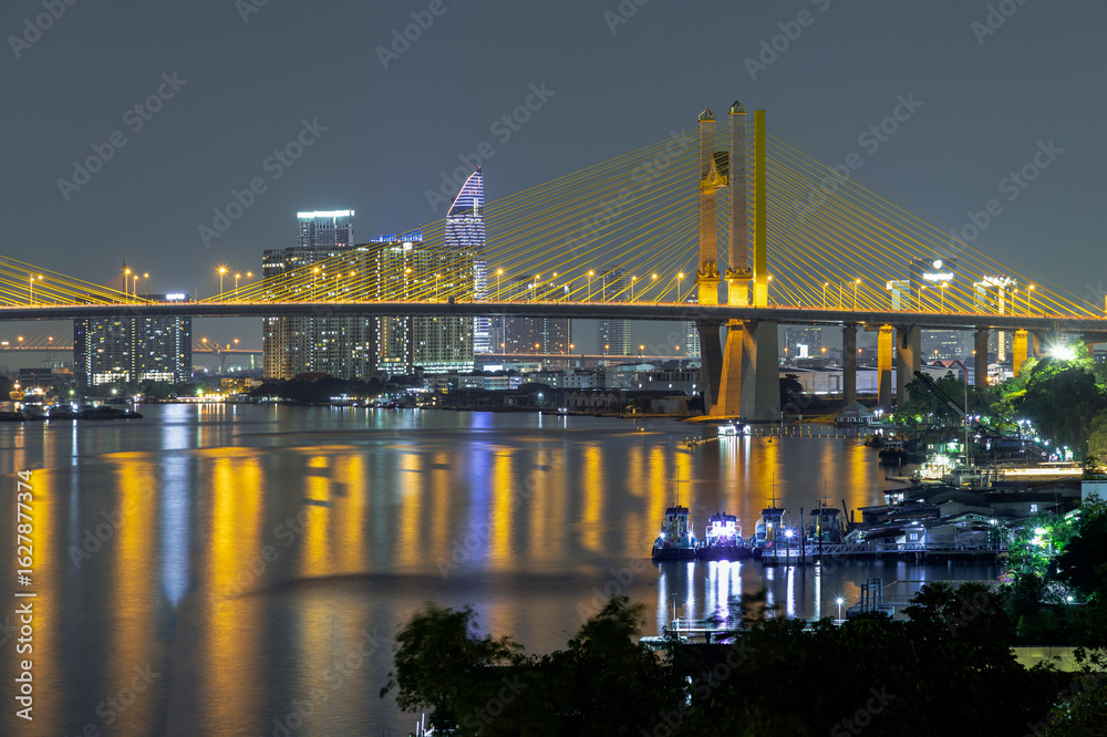 Fototapeta premium Scenery view after sunset of Thotsamarachan Bridge (Rama IX Bridge) crossing Chao phraya river with skyscraper in the background. Beautiful city of Bangkok with bright glowing lights in the sundown.