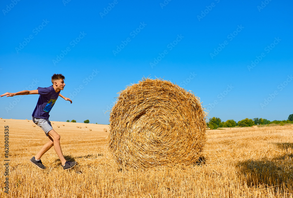 Teen boy jumping near a large round hay bale in a harvested wheat field on a sunny summer day. Fun rural activity in the countryside with blue sky and golden landscape background Teen boy jumping near a large round hay bale in a harvested wheat field on a sunny summer day. Fun rural activity in the countryside with blue sky and golden landscape background