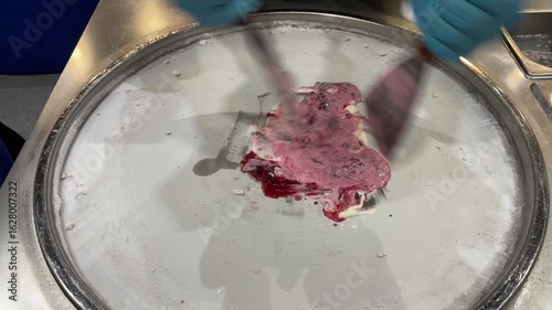 Street vendor preparing fresh rolled ice cream on a cold pan using metal spatulas, mixing creamy base with chocolate, red berry sauce and toppings