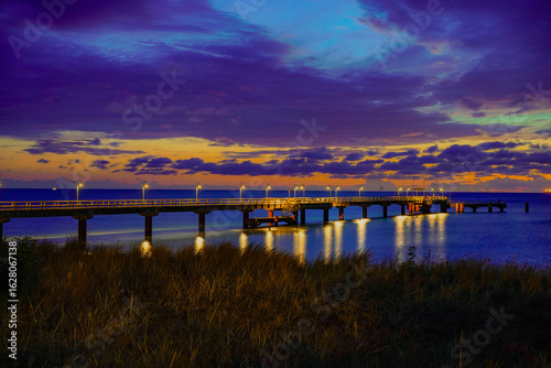 Fototapeta Naklejka Na Ścianę i Meble -  An illuminated pier extends into the Baltic Sea, the lights reflecting off the water under a cloudy morning sky. Sun and clouds at dawn on the Baltic Sea beach of 18586 Göhren, Rügen Island