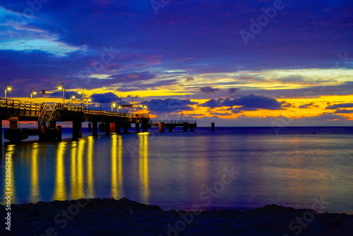 Fototapeta Naklejka Na Ścianę i Meble -  A long pier illuminates the calm ocean during the dramatic sunrise, bathed in golden and blue hues. Sun and clouds at dawn on the Baltic Sea beach of 18586 Göhren, Rügen Island