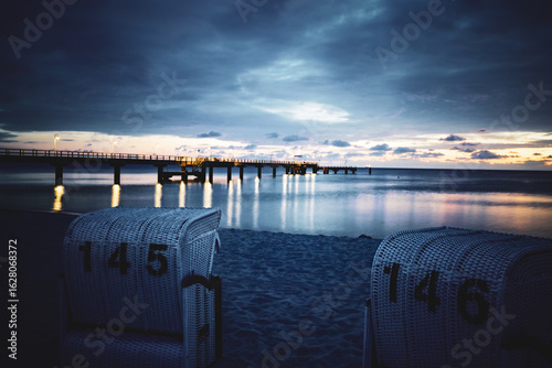 Fototapeta Naklejka Na Ścianę i Meble -  A quiet morning on the beach with an illuminated pier, framed by empty beach chairs and an atmospheric sky. Sun and clouds at dawn on the Baltic Sea beach of 18586 Göhren, Rügen Island