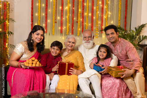 Indian happy joint family celebrating Diwali festival with full thali of laddu and gifts on the occasion of Diwali season in colorful decorated home background - Diwali holidays festive