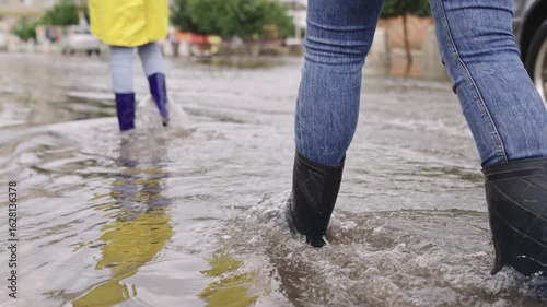 girls in raincoats and rubber boots walk along road flooded with torrential rains, their feet walk through puddles city, splashing water to the sides, the flood is on street, car is driving on water.