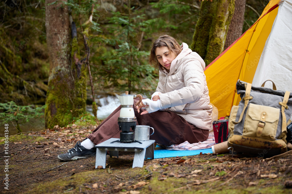 Fototapeta premium Hiker woman preparing coffee outdoor near the camping tent. A female traveler in the forest in early spring holds hot coffee in a tourist mug.