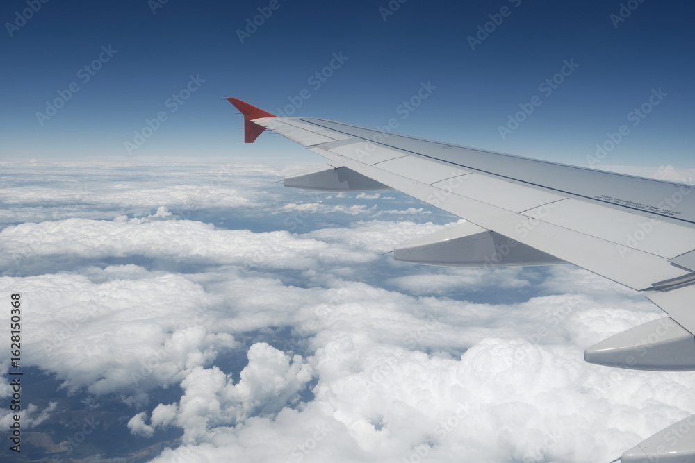 Obraz premium A peaceful in-flight view from a passenger seat showing an aircraft wing soaring above soft clouds under a bright blue sky.