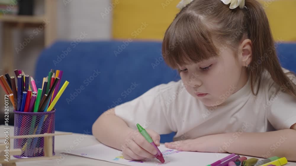 Young girl is drawing with crayons on a piece of paper. She is using a green marker and is focused on her drawing. The scene is set in a room with a couch and a bookshelf Young girl is drawing with crayons on a piece of paper. She is using a green marker and is focused on her drawing. The scene is set in a room with a couch and a bookshelf