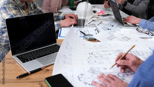 Close-up of a civil engineer working on the drawings of a modern business center. A group of people in the office are discussing profitability of project