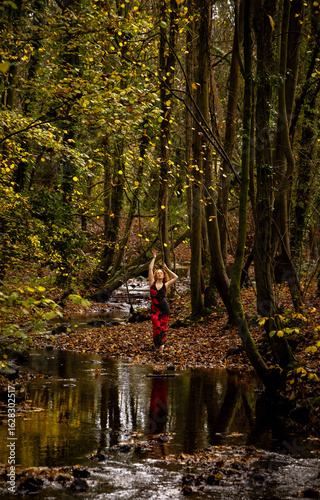 Beautiful red-haired model, woman, lady, wearing a poppy dress, reaching up into a Cathedral of autumn trees, all reflected into the mirror of a flowing river 