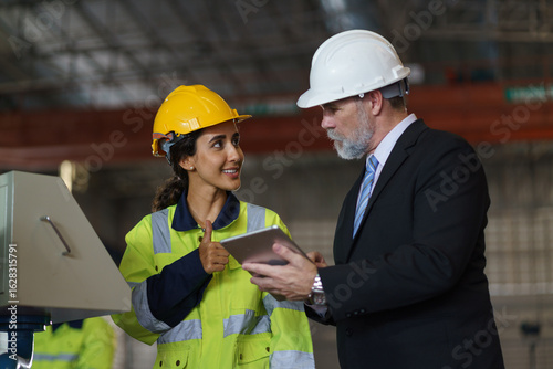 A foreman and a woman worker are standing in a factory, looking at a tablet. The engineer talks with the woman's workers. They wear a yellow and white safety helmet for safety.