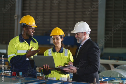 The workers are standing in a factory, looking at a tablet. The engineer talks with the woman's workers. They wear a yellow and white safety helmet for safety.