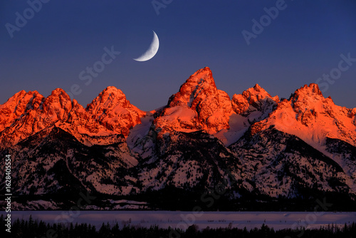 Sunrise on Teton Mountain Range in Wyoming Alpen Glow Orange and Pink on Rugged Mountains with Crescent Moon