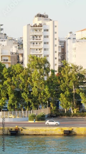 Wallpaper Mural Morning cityscape in Piraeus, Athens, Greece. Green trees in the foreground, light-toned apartment buildings in the background. Visible street with parked car, passing motorcycle, and partial sea view Torontodigital.ca