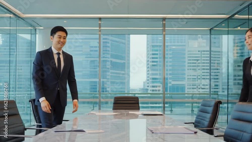 Two asian businesspeople shaking hands after reaching an agreement on a deal, standing on opposite sides of a meeting table in a modern office with cityscape view