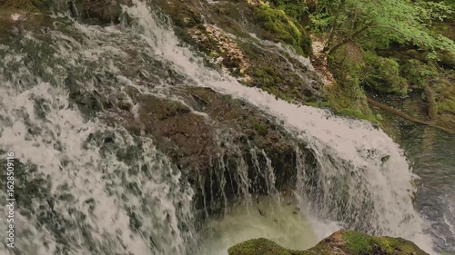 A tranquil forest creek winds through the trees with gentle sunlight filtering through the leaves. In the background, hikers stroll along a woodland path near the reflective water, capturing the calm 