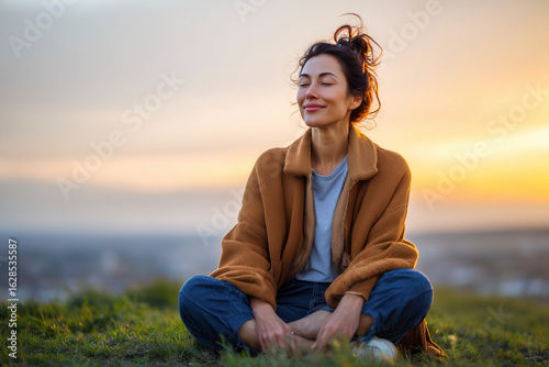 Young woman meditating outdoors at sunset, enjoying peace and mindfulness while sitting cross-legged on grass