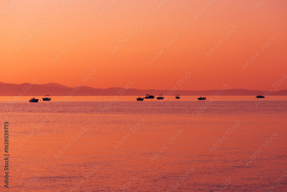 Fototapeta premium Silhouetted boats floating on calm sea during vibrant orange sunrise with distant mountains and smooth water reflections.
