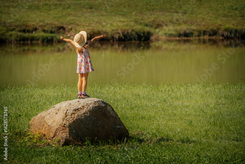 Vászonkép A young girl in a floral dress and sunhat stands on a large rock by a pond, arms outstretched in joy