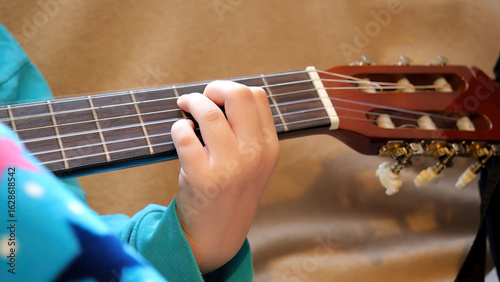 Child's hand pressing strings on classical acoustic guitar neck during home practice, closeup view of fingers forming a chord on wooden fretboard, music education and instrument learning concept