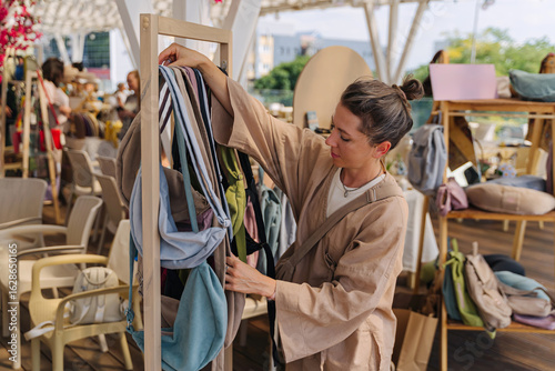Woman Artisan Arranging Handmade Eco-Friendly Bags and Backpacks at Outdoor Craft Market. Artisanal Goods, Ceramics, Candles, textile at Summer Outdoor Craft Market
