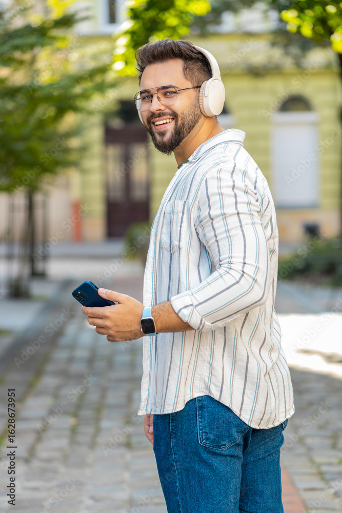 Fototapeta premium Back view of carefree young man tourist in wireless headphones listening favorite energetic disco music dancing outdoors. Caucasian guy traveler walking spinning on urban city street. Town lifestyle.