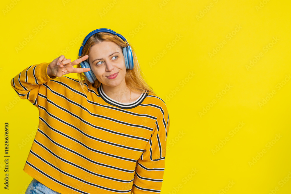 Fototapeta premium Happy joyful Caucasian woman in blouse listening music on headphones and dancing disco fooling around having fun expressive gesticulating hands. Young adult girl isolated on yellow background indoors