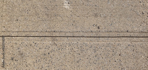 Close-up of a concrete sidewalk surface with visible aggregate and straight expansion joint lines. Detailed urban texture showing grit, small stones, and rough cement finish