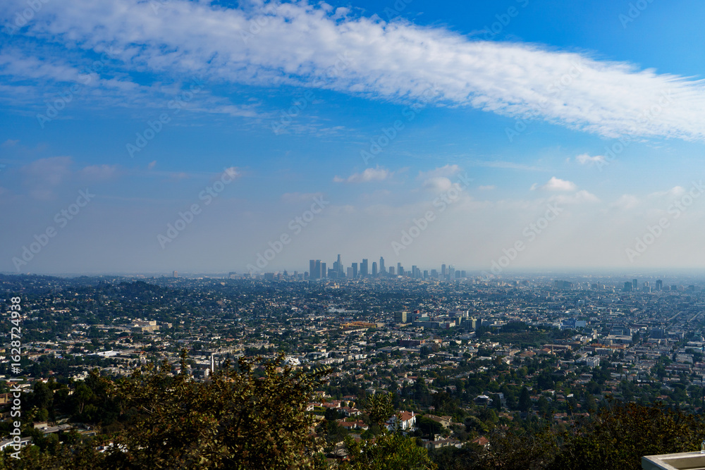 Fototapeta premium A high-angle, panoramic view captures the sprawling Los Angeles cityscape, with the downtown skyline rising in the distance beneath a bright blue, cloud-strewn sky.