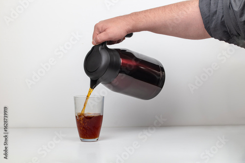 Man pouring hot coffee from black thermos bottle into glass cup on white background, morning beverage drink preparation concept