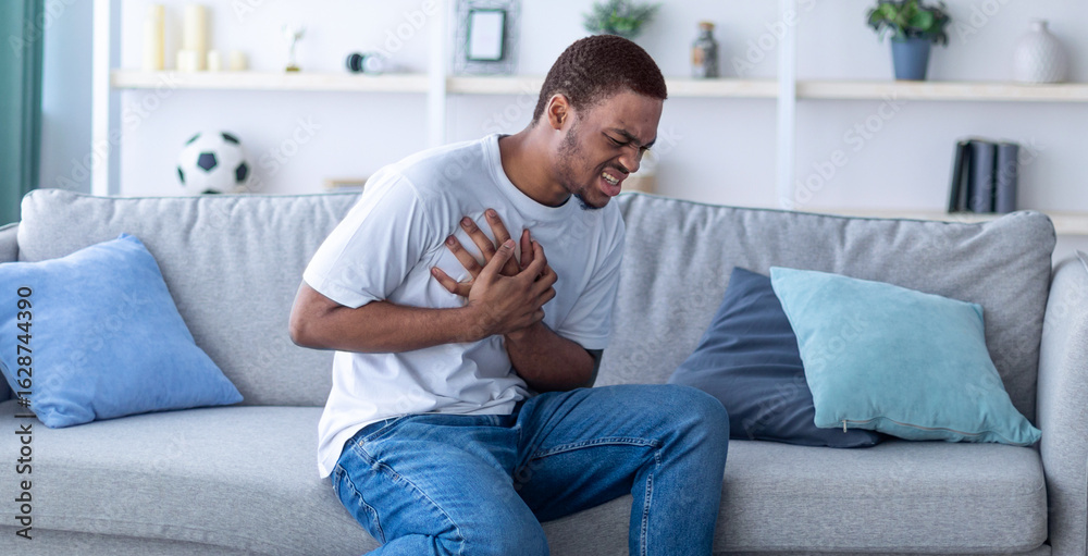 Millennial African American guy having heart attack, touching his chest in pain, sitting on sofa at home. Young black man suffering from cardiovascular ache, getting infarction indoors Millennial African American guy having heart attack, touching his chest in pain, sitting on sofa at home. Young black man suffering from cardiovascular ache, getting infarction indoors