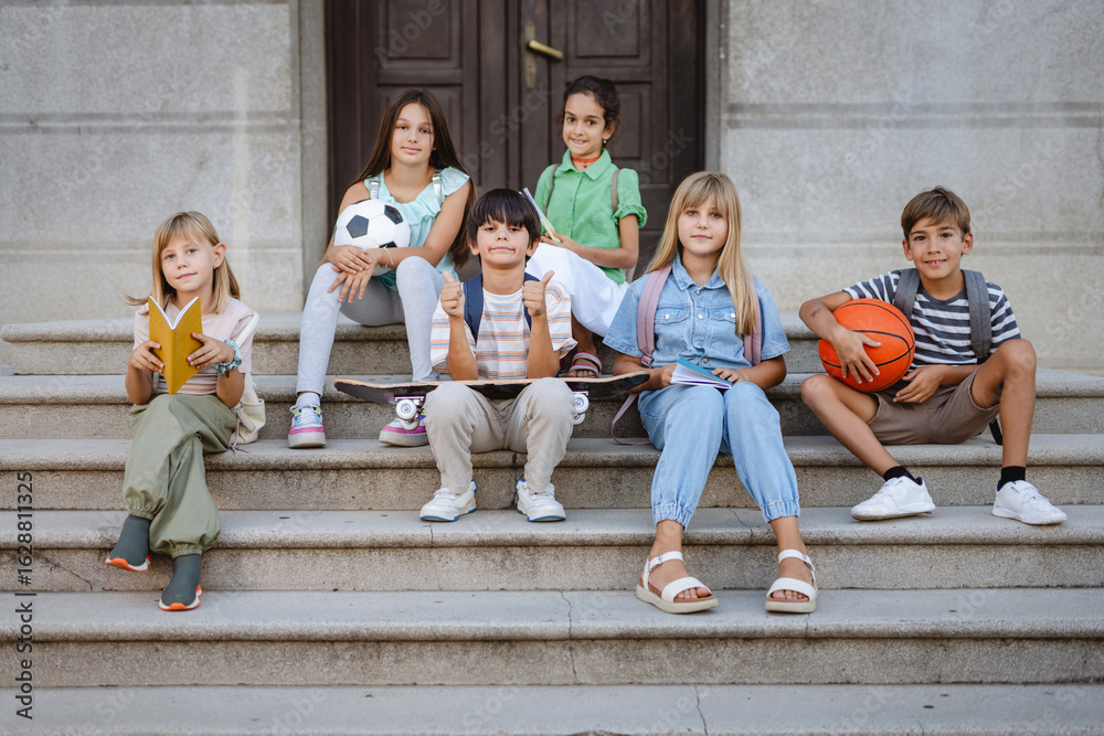 Fototapeta premium Students sitting on school steps with sport and study items showing thumbs up