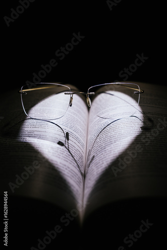 Open book illuminated with heart-shaped light and glasses on a black background.