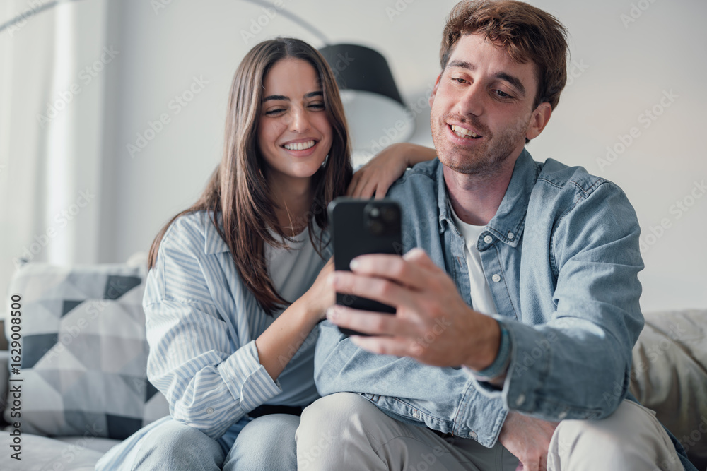Fototapeta premium Cute couple greeting having fun video calling long distance friends and family catching up. Young man holding phone with girlfriend sitting on the sofa laughing and enjoying technology