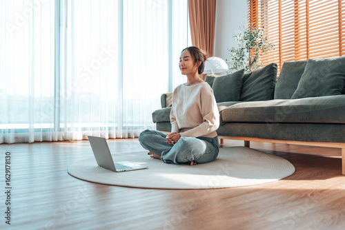 Happy young Asian woman practicing yoga and meditation at home sitting on floor in living room in lotus position with laptop