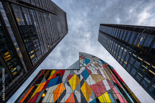 Low-angle shot of two modern skyscrapers, one featuring a vibrant, colorful, geometric facade, juxtaposed against a darker, more traditional skyscraper.