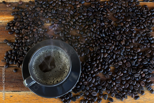 A cup of hot coffee is placed on a wooden table, with coffee beans, top view.