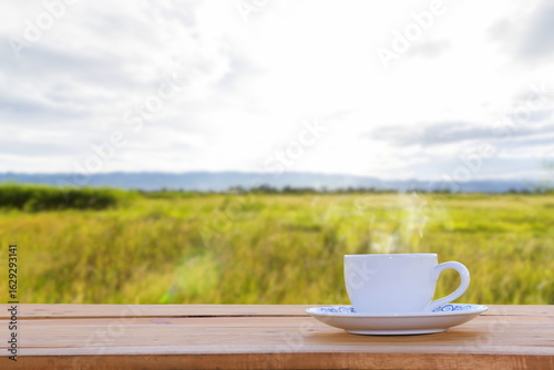 A cup of hot coffee is placed on a wooden table, overlooking the landscape, with blurred green fields, mountains and sky in the background.