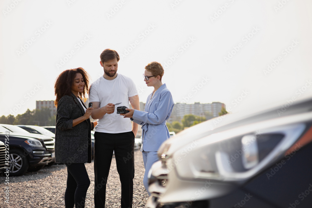 Obraz premium A couple is paying for a car at a dealership using a credit card, assisted by a saleswoman in a car lot.