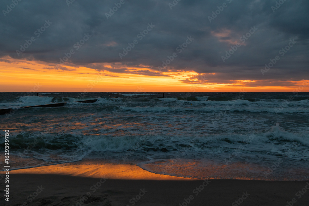 Fototapeta premium Dramatic sunset over a stormy sea with dark clouds, golden-orange horizon, and waves crashing onto the sandy shore, reflecting the warm colors of the sky.