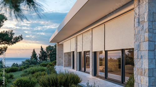 Modern stone villa with closed outdoor blinds at sunset. Luxury coastal home with privacy and stunning sea view.