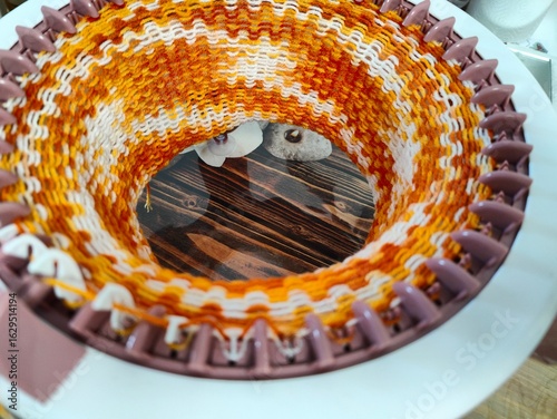 Circular knitting machine with multicolor yarn. Close-up view of a round knitting loom in action, weaving orange, white, and yellow yarn into a tubular knitted fabric
