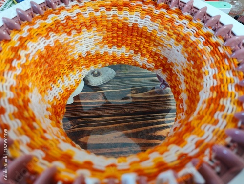 Circular loom knitting with orange yarn. Close-up view of a circular loom knitting project using orange and white yarn, showing pattern and crafting process details