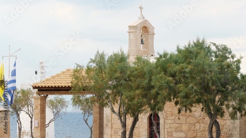 Wallpaper Mural Partial view of a small stone church with terracotta roof and cross-topped bell tower near the sea, framed by trees and flags, with calm blue water and clear sky in the background Torontodigital.ca