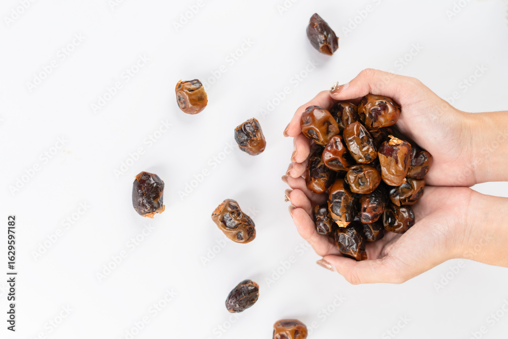 Obraz premium A pair of hands gently holding a bunch of dried dates over a clean white surface, with scattered dates adding a rustic and natural touch.