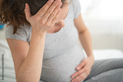 Expecting mother holding her head in pain while resting on bed. Difficult moment of pregnancy migraine and maternal health concern at home.