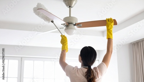 A woman in yellow gloves carefully cleans a ceiling fan with a duster in a bright room.