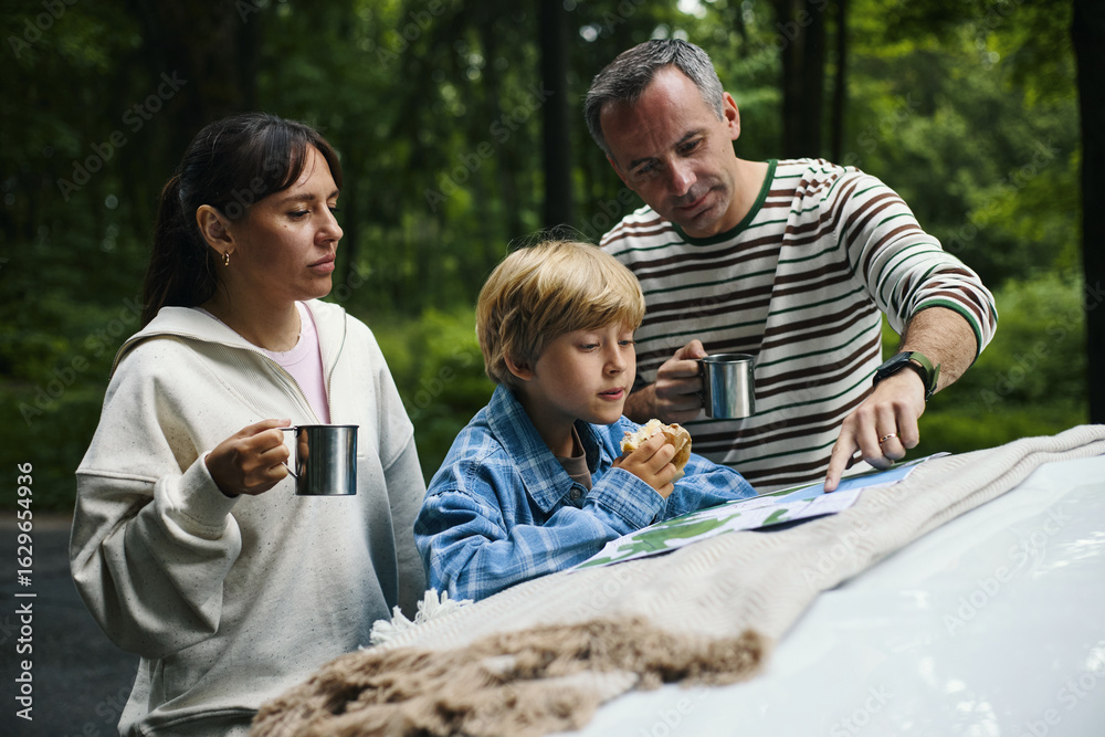 Caucasian middle aged man and woman standing beside Caucasian child eating snack and holding mug, man pointing at map on car hood during family road trip in forest Caucasian middle aged man and woman standing beside Caucasian child eating snack and holding mug, man pointing at map on car hood during family road trip in forest