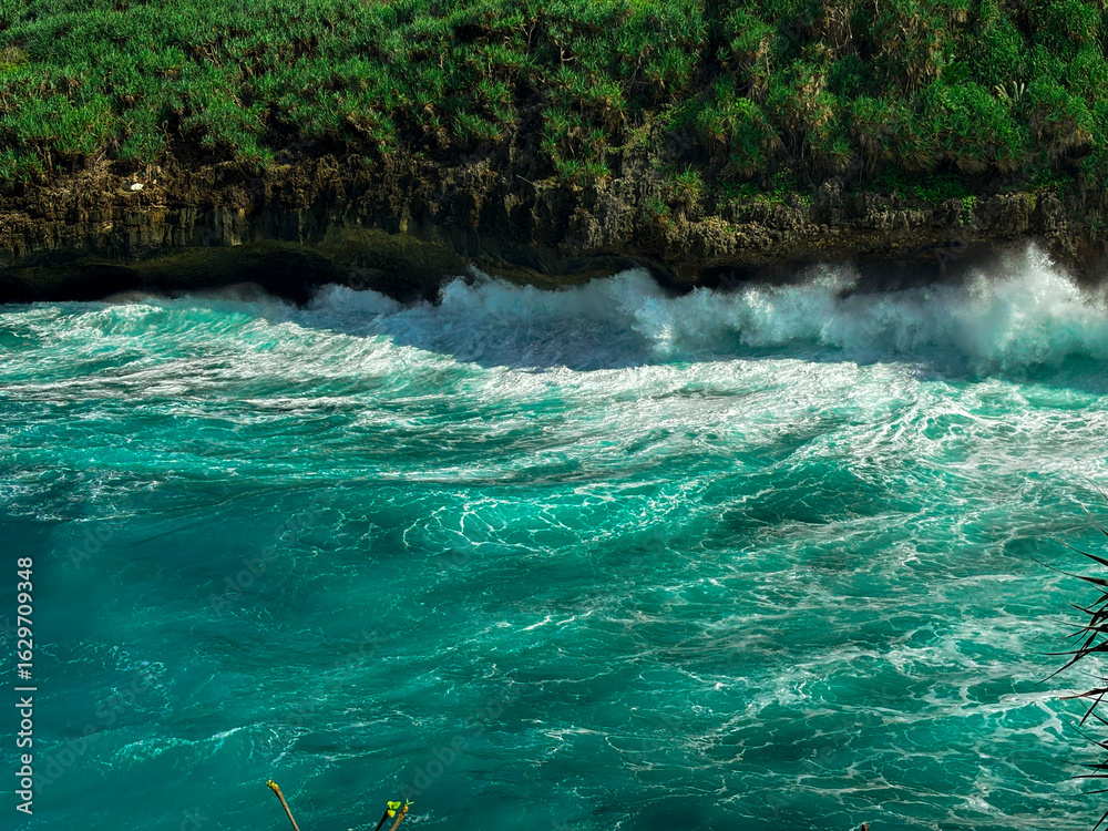 Naklejka premium Turquoise Sea Waves: Captivating turquoise waves crash against the rugged coastline. Lush greenery frames the stunning view, showcasing the raw power and beauty of the ocean. 