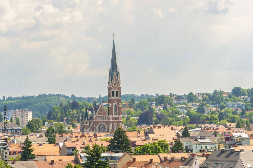 View of Herz Jesu Kirche soaring above a tapestry of red-tiled roofs, nestled amidst lush greenery under a soft sky, Herz jesu kirche church, Graz, Austria. View of Herz Jesu Kirche soaring above a tapestry of red-tiled roofs, nestled amidst lush greenery under a soft sky, Herz jesu kirche church, Graz, Austria.